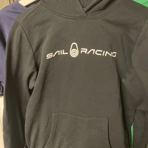 Svart hoodie från Sail Racing - Säljer en svart hoodie från Sail Racing med vit logga på bröstet och en mindre logga på huvan. Tröjan har en klassisk känguruficka framtill och långa ärmar. 