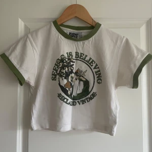 Vit och grön t-shirt från Reclaimed Vintage - Säljer en cool vit t-shirt från Reclaimed Vintage med gröna detaljer och texten 'Seeing is Believing'. Den har korta ärmar och en croppad passform. Perfekt för en avslappnad stil.