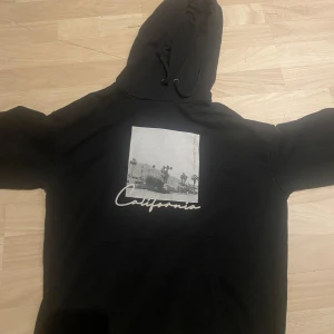 Svart hoodie-california - Säljer en svart hoodie med ett tryck av palmer och texten 'California' på framsidan. Tröjan har en klassisk design med huva och långa ärmar. Perfekt för en avslappnad stil.