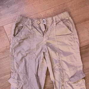 Beige cargobyxor från BDG - Säljer ett par beige cargobyxor från BDG. Byxorna har flera praktiska fickor och en avslappnad passform. Perfekta för en casual look med en touch av stil. De har en elastisk midja för extra komfort.