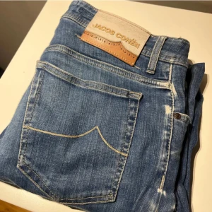 Blå jeans från Jacob Cohën - Snygga blå jeans från Jacob Cohën med klassisk femficksdesign och en läderpatch med logotyp på baksidan. Jeansen har en rak passform och är tillverkade i ett slitstarkt denimtyg. Perfekta för en stilren look. Modellen är bard fast och storleken är W33.
