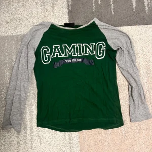 Grön och grå långärmad tröja med text - Säljer en grön och grå långärmad tröja med texten 'GAMING YOU VS. ME' på framsidan. Perfekt för gaming-entusiaster! Tröjan har en bekväm passform och är gjord i mjukt material.