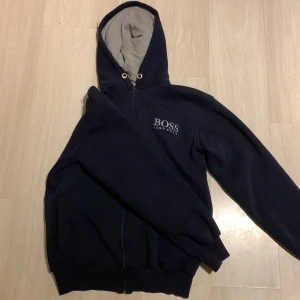 Svart hoodie från Hugo Boss - Säljer en stilren svart hoodie från Hugo Boss med dragkedja och huva. Den har en broderad logga på bröstet och är tillverkad i ett mjukt material. Perfekt för en avslappnad look.
