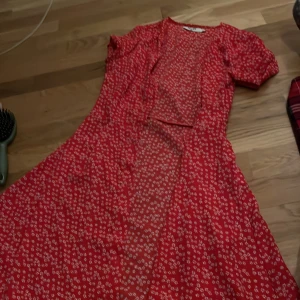 Röd blommig klänning - Charmig röd klänning med ett sött blommönster. Den har korta ärmar och en luftig passform som ger en härlig sommarkänsla. Perfekt för varma dagar och avslappnade tillfällen.