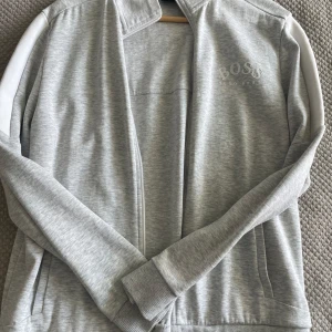 Grå zip-tröja från Hugo Boss - Snygg grå zip-tröja från Hugo Boss med vit logga på bröstet. Tröjan har långa ärmar och ribbade muddar. Nyskick