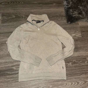Beige tröja med dragkedja från jack & Jones  - Säljer en stilren beige tröja från jack & Jones med dragkedja vid halsen. Tröjan har långa ärmar och ribbade muddar vid ärmslut och nederkant. Perfekt för kyligare dagar!