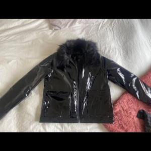 Svart glansig jacka med fuskpäls - Snygg svart glansig jacka med fuskpäls på kragen. Jackan har en stilren design med dragkedja framtill och långa ärmar. Perfekt för att ge en edgy touch till din outfit. 🖤