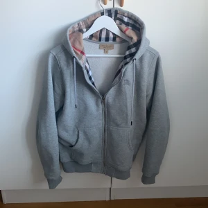 Grå hoodie från Burberry - Säljer en stilren grå hoodie från Burberry med klassiskt rutigt mönster på insidan av huvan. Tröjan har dragkedja framtill och ribbade muddar vid ärmslut och nederkant. Perfekt för en avslappnad och trendig look.