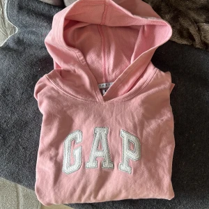 Rosa hoodie från GAP - Säljer en rosa hoodie från GAP med klassisk logga på bröstet. Tröjan har en känguruficka och långa ärmar. Obs, små fläckar på båda ärmarna, därav det låga priset. 
