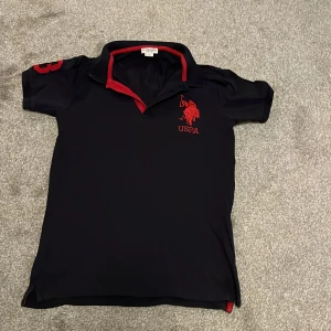 Mörkblå pikétröja från U.S. Polo Assn. - Snygg mörkblå pikétröja från U.S. Polo Assn. med röda detaljer. Tröjan har korta ärmar och ett stort rött nummer '3' på ärmen. Märkets logga är broderad i rött på bröstet. Perfekt för en sportig look! Står för 14 år men är som xs/s skick: 9/10 knappt använd 