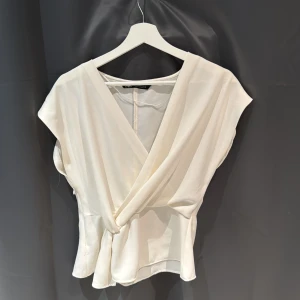 Vit omlottopp - Säljer en stilren vit omlottopp från Zara. Toppen har en v-ringad design och är ärmlös, vilket ger en elegant look. Perfekt för både vardag och festliga tillfällen.