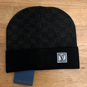 Svart mössa från Louis Vuitton - Snygg svart mössa från Louis Vuitton med ett diskret mönster och en broderad LV-logga framtill. Perfekt för att hålla sig varm med stil. Skriv vid intresse