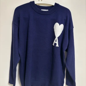 Mörkblå stickad tröja från Ami - Säljer en snygg mörkblå stickad tröja från Ami med ett vitt hjärta och bokstaven 'A' på bröstet. Tröjan har långa ärmar och en rund halsringning. Perfekt för kyliga dagar!