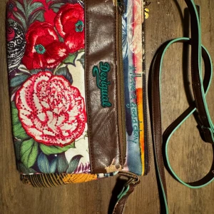 Färgglad axelväska från Desigual - Säljer en unik och färgglad axelväska från Desigual med blommigt mönster och randiga detaljer. Väskan har en justerbar axelrem i brunt skinn och en praktisk dragkedja. Perfekt för att ge din outfit en färgklick! Den är i använt skick! 