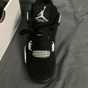 Jordan 4s  - Snygga svarta Nike Air Jordans med vita detaljer och klassisk snörning. Skorna har en ikonisk design med nätmönster på sidorna och Jumpman-loggan på plösen. Perfekta för dig som vill ha en stilren och sportig look. Köpt från stock x mer använda än på bilderna men fortfarande fina och fräscha 