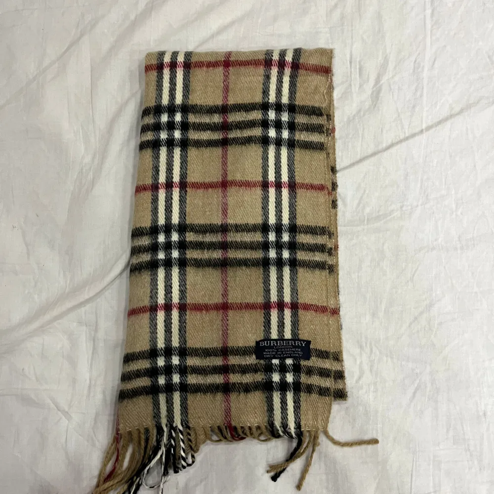 Säljer en klassisk beige rutig halsduk från Burberry i 100% kashmir. Den har ett tidlöst rutmönster i svart, rött och vitt. Perfekt för att hålla dig varm och stilren under kyliga dagar. Tillverkad i England. . Asusteet.