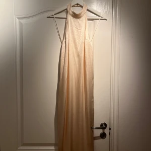 Champagnefärgad långklänning från Zara - Elegant champagnefärgad helt ny långklänning från Zara med halterneck-design och öppen rygg. Klänningen har en mjuk och glansig yta som ger en lyxig känsla. Perfekt för speciella tillfällen där du vill glänsa lite extra. 💫