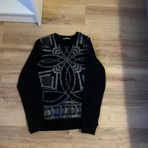 Svart tröja med geometriskt mönster versacejeans - Snygg svart tröja med ett unikt geometriskt mönster i grått. Tröjan har en v-ringad hals och långa ärmar, perfekt för en stilren look. Passar bra till både jeans och chinos.