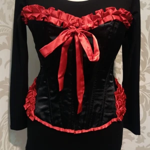Svart korsett med röda detaljer storlek XL - Elegant svart korsett med röda rosetter och volangdetaljer. Den har en åtsittande passform och är perfekt för en festlig look. Tillverkad i ett glansigt material som ger en lyxig känsla. Ej underklädeskorsett. Storlek XL.