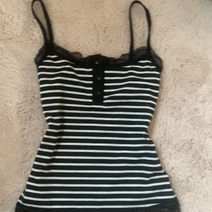Svartvit randig linnetopp med spetsdetaljer - Säljer en snygg svartvit randig linnetopp med spetsdetaljer vid halsringningen. Toppen har smala axelband och knappar framtill. 