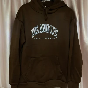 Brun hoodie med Los Angeles-tryck - Säljer en brun hoodie med texten 'Los Angeles California' tryckt på framsidan. Tröjan har en klassisk känguruficka och en justerbar huva med dragsko. Perfekt för en avslappnad stil.