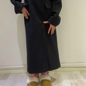 Svart lång kappa - Elegant svart lång kappa med klassisk design. Perfekt för kyligare dagar med sin stilrena look och långa ärmar. Kappan har en rak passform och dolda knappar framtill.