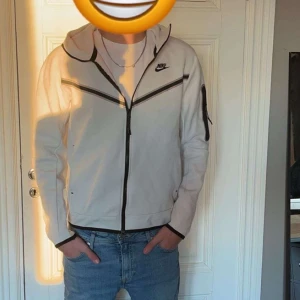 Nike tech - Säljer en stilren vit hoodie från Nike med dragkedja och huva. Jackan har svarta detaljer och en liten Nike-logga på bröstet. Perfekt för en avslappnad look.