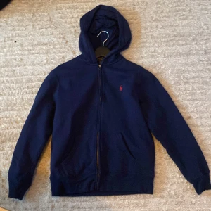 Mörkblå hoodie från Ralph Lauren(158 cm) - Säljer en snygg mörkblå hoodie från Ralph Lauren med dragkedja och det klassiska röda logotypen på bröstet. 14-16 år, liten i storleken så ungefär 158 cm. Perfekt för en avslappnad stil. Den har en praktisk huva och långa ärmar.