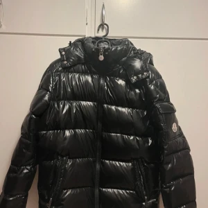Svart dunjacka från Moncler - Säljer en snygg svart dunjacka från Moncler. Jackan har en glansig finish och är utrustad med dragkedja och huva. Perfekt för kyliga dagar med sin varma och stilrena design. Moncler-loggan syns tydligt på ärmen. jag vill bara få jackan såld så snabbt som möjligt därför är priset så lågt