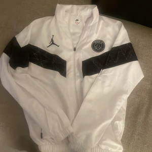 Vit vindjacka från Jordan x PSG - Säljer en stilren vit vindjacka från Jordan i samarbete med Paris Saint-Germain. Jackan har svarta detaljer och logotyper på bröstet och ärmarna. Den är utrustad med dragkedja och har en hög krage för extra stilpoäng. Perfekt för dig som vill ha en sportig look!