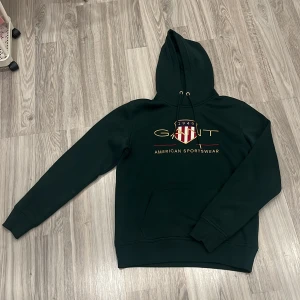 Mörkgrön hoodie från GANT - Säljer en snygg mörkgrön hoodie från GANT med deras klassiska logga och texten 'American Sportswear' på framsidan. Tröjan har en stor ficka framtill och justerbar huva med snören. Perfekt för en avslappnad stil.