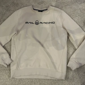 Beige sweatshirt från Sail Racing - Säljer en stilren beige sweatshirt från Sail Racing med svart logotyp på bröstet. Tröjan har långa ärmar och rund halsringning, perfekt för en avslappnad stil. Passar bra till både jeans och chinos.
