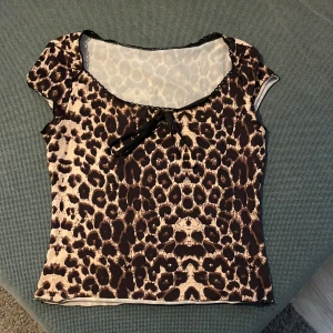 Leopardmönstrad topp med rosett - Snygg leopardmönstrad topp med korta ärmar och en söt rosett framtill. Perfekt för att ge din outfit en vild touch. Toppen har en rund halsringning och är tillverkad i ett mjukt material.