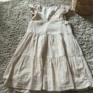 Beige klänning med volangdetaljer - Charmig beige klänning med volangdetaljer på axlarna och en v-ringad design. Klänningen har en luftig passform och är tillverkad i ett lätt material, perfekt för varmare dagar. Gjord av linne. 