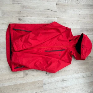 Röd vindjacka från Oakley - Säljer en snygg röd vindjacka från Oakley med huva och dragkedja. Jackan har svarta detaljer och flera praktiska fickor med dragkedjor. Perfekt för blåsiga dagar och utomhusaktiviteter.