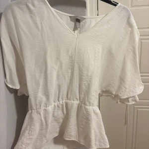 Vit blus från H&M - Säljer en söt vit blus från H&M med v-ringning och korta ärmar. Blusen har en volangdetalj i nederkant och knytband på axlarna för en justerbar passform.  Använd en gång så i mycket fint skick🩷passar även S