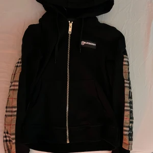 Svart hoodie från Burberry - Säljer en snygg svart hoodie från Burberry med dragkedja och klassiskt rutigt mönster på ärmarna. Den har en huva och Burberry-loggan på bröstet. Perfekt för en stilren look.