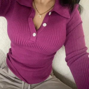 Lila ribbad tröja med knappar - Snygg lila ribbad tröja med tre glittriga knappar framtill. Tröjan har en krage och långa ärmar, perfekt för en stilren look.