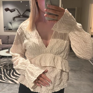 Beige blus med volangdetaljer - Säljer en stilren beige blus med volangdetaljer och v-ringning. Blusen har långa ärmar och ett lätt skrynklat material 