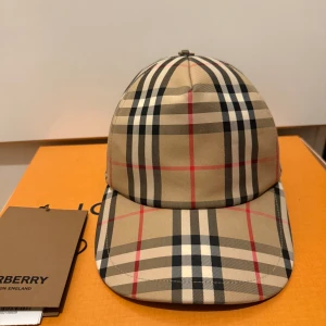 Burberry keps - Snygg rutig keps från Burberry i klassiskt beige, svart och rött mönster. Kepsen har en justerbar rem baktill och en broderad logotyp. Perfekt för att ge din outfit en stilren touch.