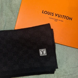 Svart halsduk från Louis Vuitton - Elegant svart halsduk från Louis Vuitton med diskret LV-logga. Tillverkad i en lyxig blandning av 60% silke och 40% ull, vilket ger en mjuk och varm känsla. Perfekt för att ge en stilren touch till din outfit.  Kvitto och lådan medföljs