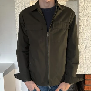 Olivgrön jacka från Threadbare - Snygg olivgrön jacka från Threadbare med dragkedja framtill och stilrena fickor på bröstet. Jackan har långa ärmar med knappar vid ärmsluten och en klassisk krage. Perfekt för en casual look. Storlek s passar m