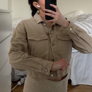 Beige jeansjacka - Säljer en snygg beige jeansjacka med knappar framtill och två bröstfickor. Jackan har en croppad design och långa ärmar, perfekt för en avslappnad stil. Passar bra till både kjol och byxor. Köpt från Ginatricot 2019. Jättefint skick