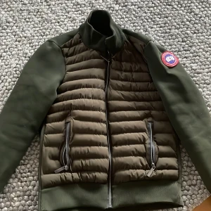 Grön jacka från Canada Goose - Säljer en snygg grön jacka från Canada Goose med quiltad design och dragkedja. Jackan har långa ärmar och en broderad logotyp på ärmen. Perfekt för kyligare dagar.