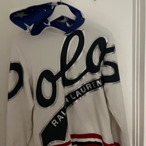 Vit hoodie från Ralph Lauren - Säljer en vit hoodie från Ralph Lauren med stor logga i svart och silver på framsidan. Hoodien har en blå insida med stjärnmönster i luvan och röd-svarta ränder vid nederkanten. Perfekt för en stilren och sportig look.