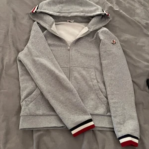 Grå hoodie från Moncler - Säljer en stilren grå hoodie från Moncler med dragkedja och luva. Tröjan har långa ärmar och ribbade muddar med röd, vit och svart rand. 