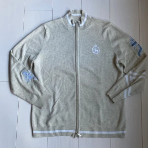 Polo Full-zip - Säljer en snygg beige full zip med tryck från Polo Club i riktigt skönt material. Tröjan har ett broderat emblem på bröstet och ett stort mönster på ryggen med texten 'Polo Club'. Den har ribbade muddar och krage med vita detaljer. Storlek S-M