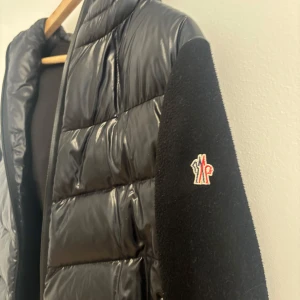 Svart dunjacka Moncler med huva - Snygg svart dunjacka från Moncler med glansigt material och huva. Jackan har långa ärmar och en dragkedja framtill. Sann i storlek! 