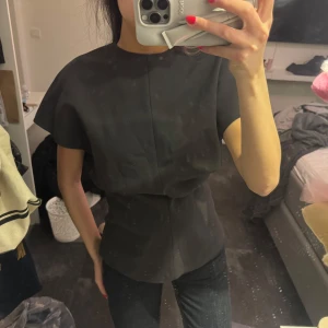 Mörkgrå topp med korta ärmar - Säljer en stilren mörkgrå topp med korta ärmar. Toppen har en elegant och enkel design som passar perfekt till olika tillfällen. Den är figursydd för en smickrande passform. Ny med prislapp 