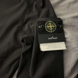 Svart vindjacka från Stone Island - Snygg svart vindjacka från Stone Island med huva och dragkedja framtill. Jackan har en stilren design med märkets ikoniska logga på ärmen. Perfekt för en modern och sportig look.